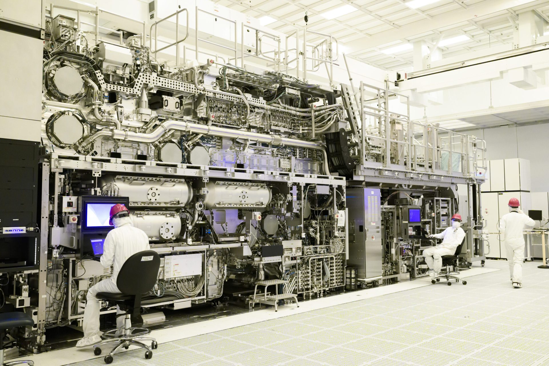 Technicians in a clean room work on an advanced ASML lithography machine with multiple screens and intricate machinery components.