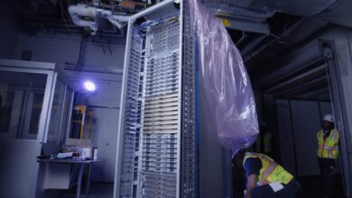 A large, vertically standing, unbranded server rack filled with equipment is being installed by workers in a dimly lit data center.
