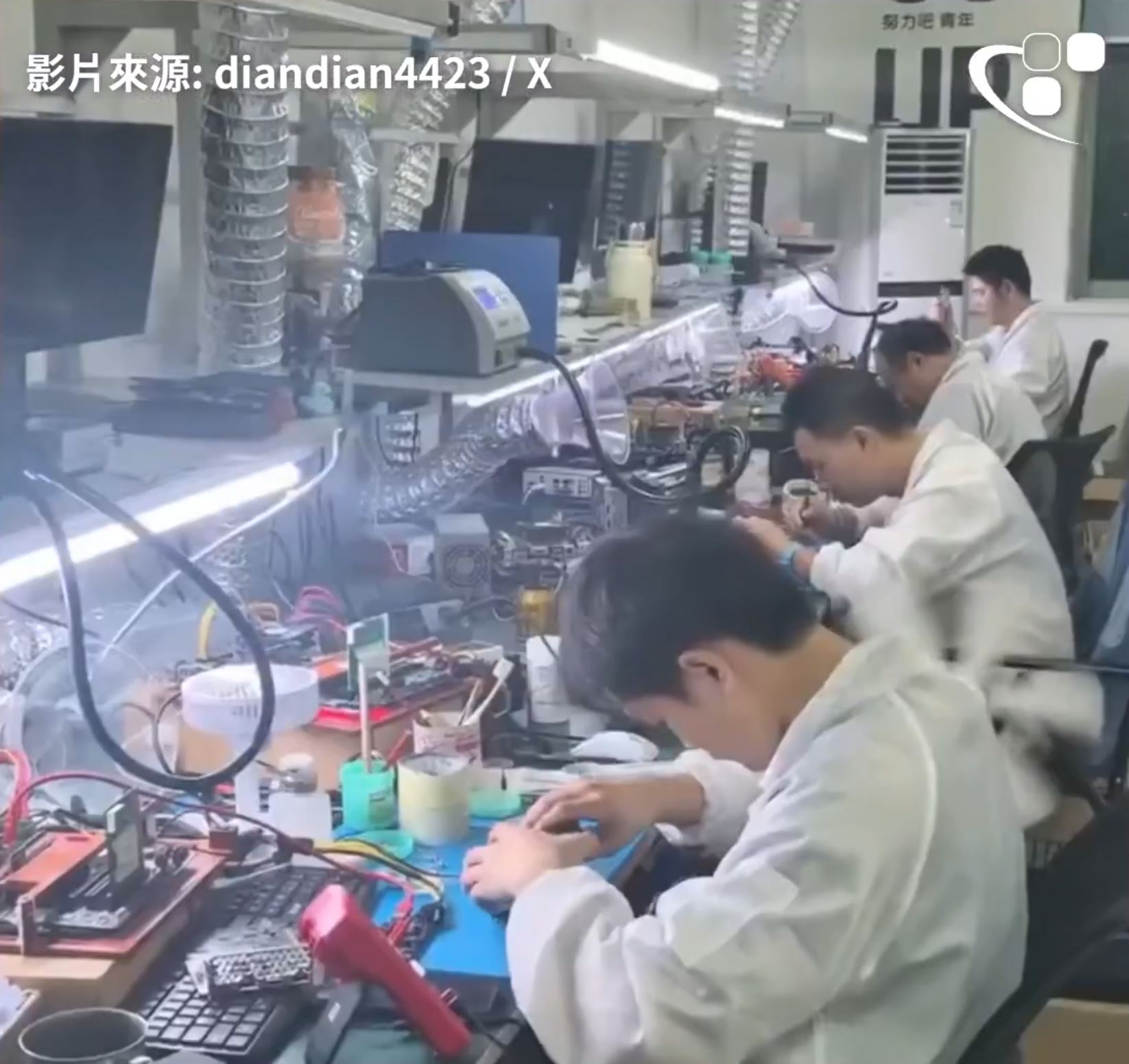 Several technicians in lab coats are working on electronics at a cluttered workstation, equipped with various tools and components, in a well-lit lab.